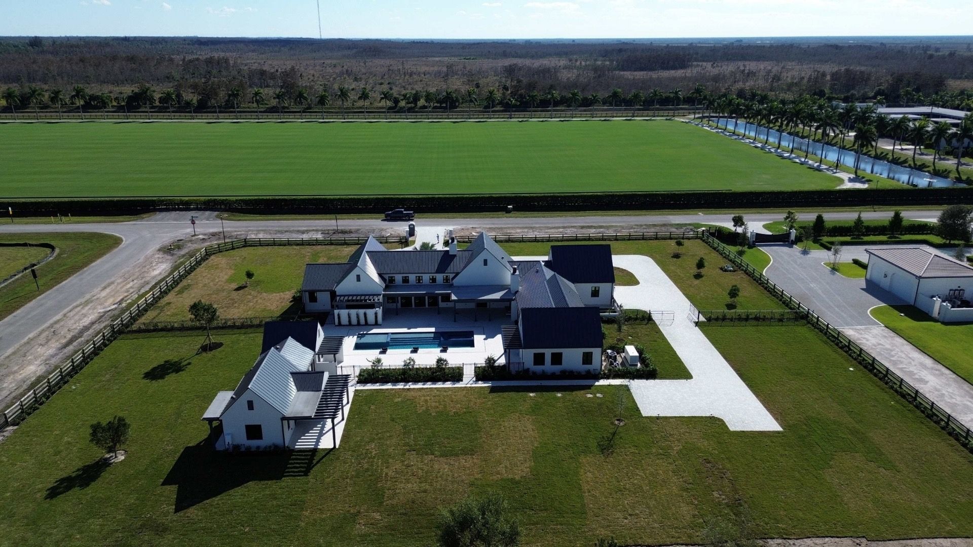 Aerial view of a large white house with a pool, surrounded by green lawns and a field, sunny day.
