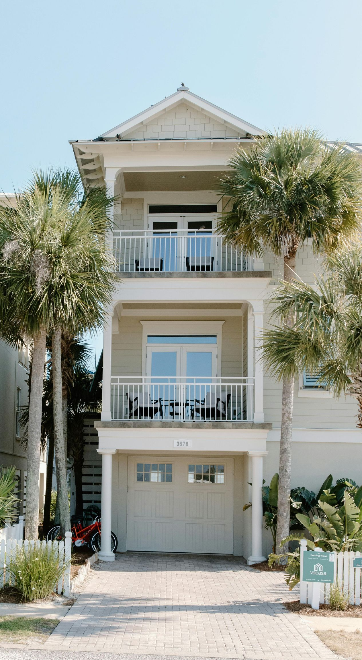 Beige multi-story house with balconies and garage, palm trees on either side, clear blue sky.