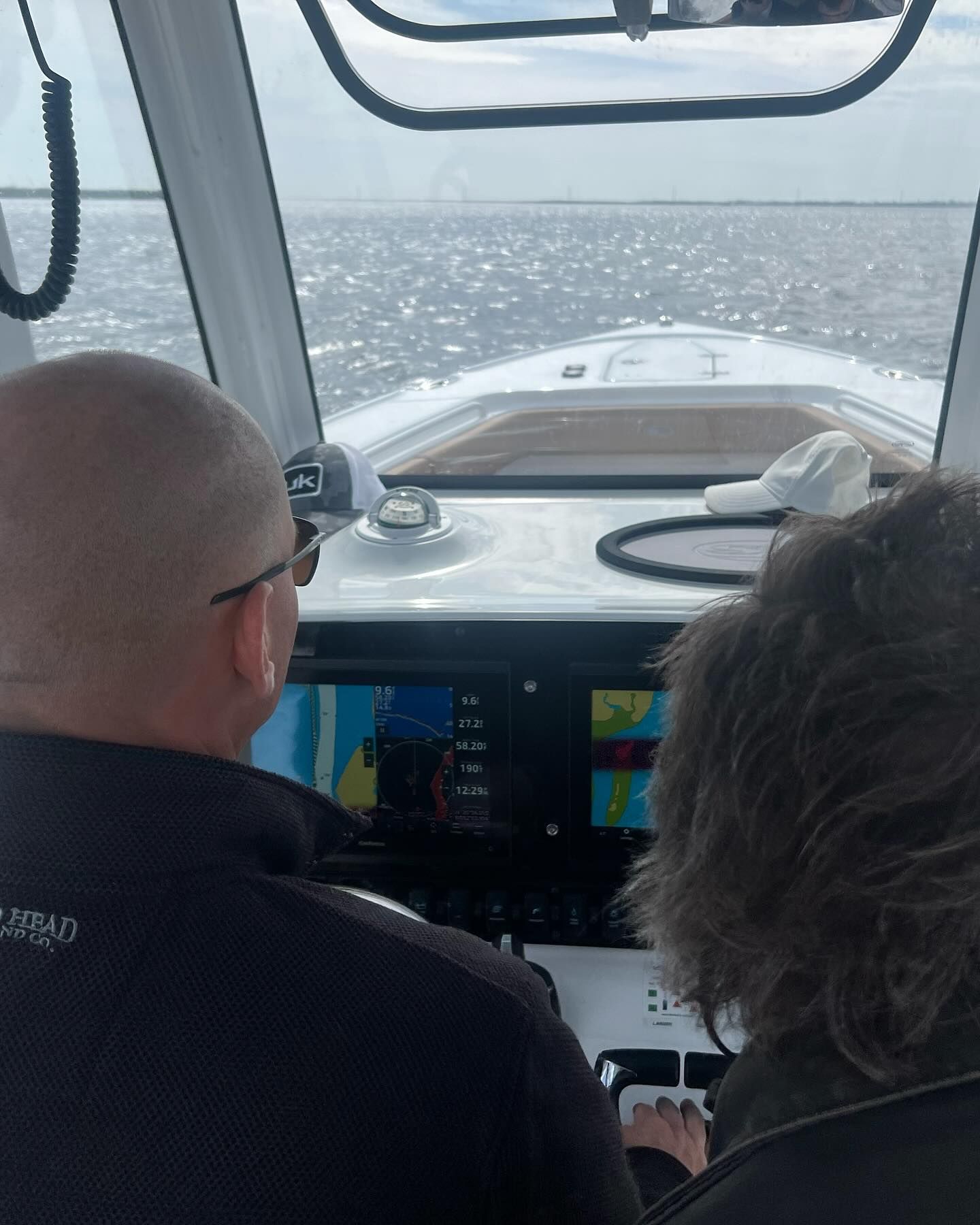 Two people driving a boat, looking ahead at the ocean. Dashboard and windshield visible.