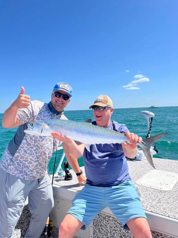 Two men on a boat holding a fish, smiling in front of the ocean under a blue sky.