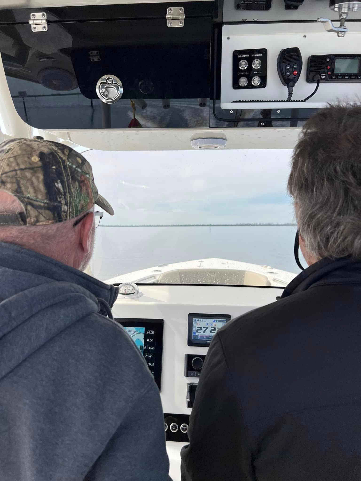 Two men in a boat, looking out at water. One wears a camo hat. Navigation instruments are visible.