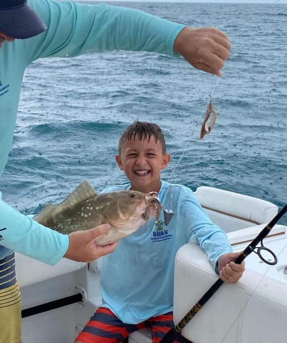 Boy smiles holding a fish on a boat, another fish on a hook above him. A man assists. Ocean setting.