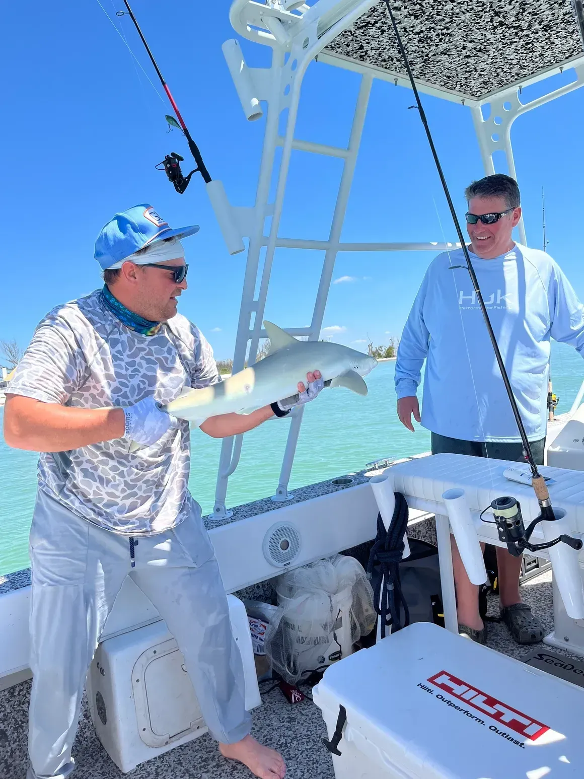 Man holding a small shark on a boat, another man smiling. Sunny day, blue water.