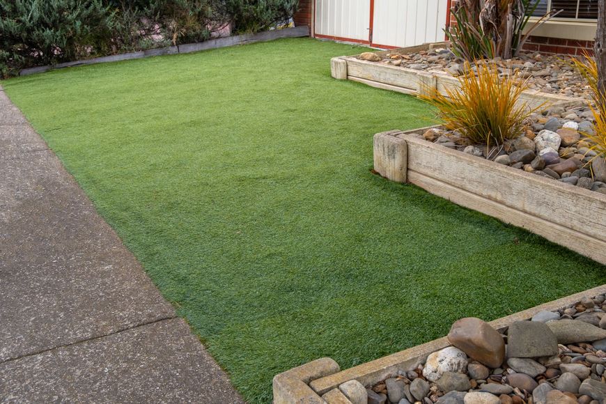 Green artificial turf lawn next to a concrete sidewalk and flower beds filled with rocks and plants.