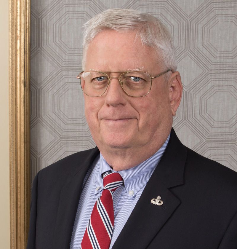 Portrait of a man in a navy suit, red striped tie, and glasses against a patterned wall