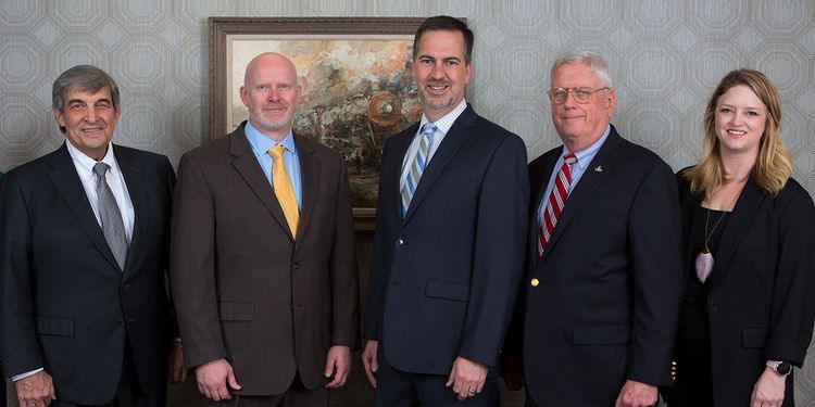 Five people in business attire posing for a formal group photo indoors.