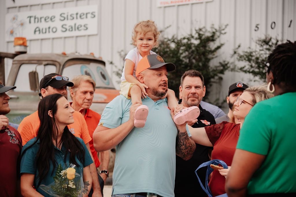 A man is carrying a little girl on his shoulders in front of a group of people. | Chuck's Garage