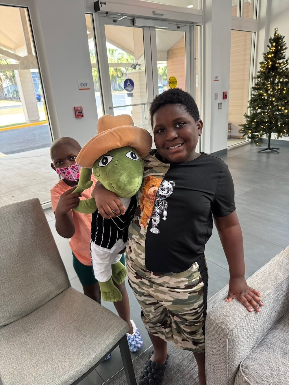 Two boys smiling, holding a stuffed frog wearing a cowboy hat and vest; indoor setting.