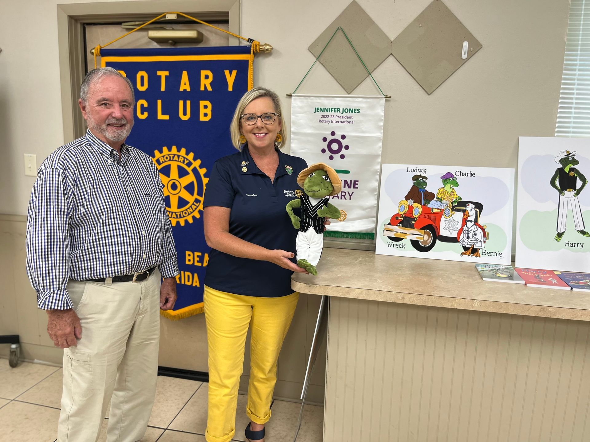 Two people stand near a Rotary Club banner and artwork. Woman holds a stuffed animal.