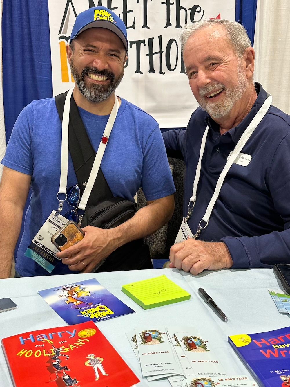 Two men smiling, posing for photo at booth. One in blue shirt/cap, one in navy shirt. Books on table.