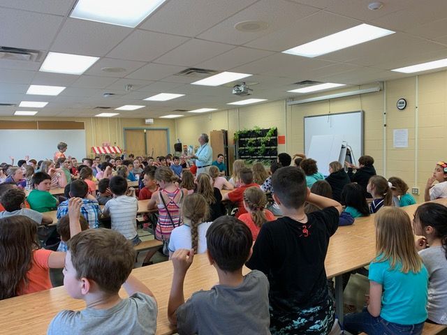 Children seated at tables, listening to a presentation in a large, bright cafeteria.