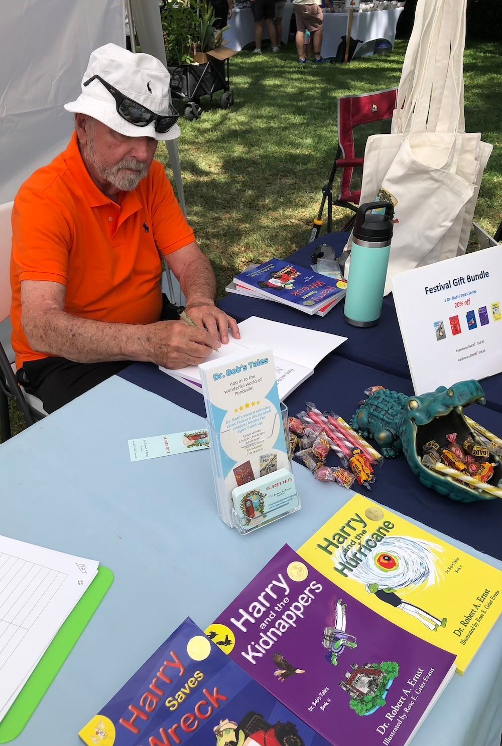 Man in orange shirt signs books at a fair. Books and merchandise on the table, outdoor setting.