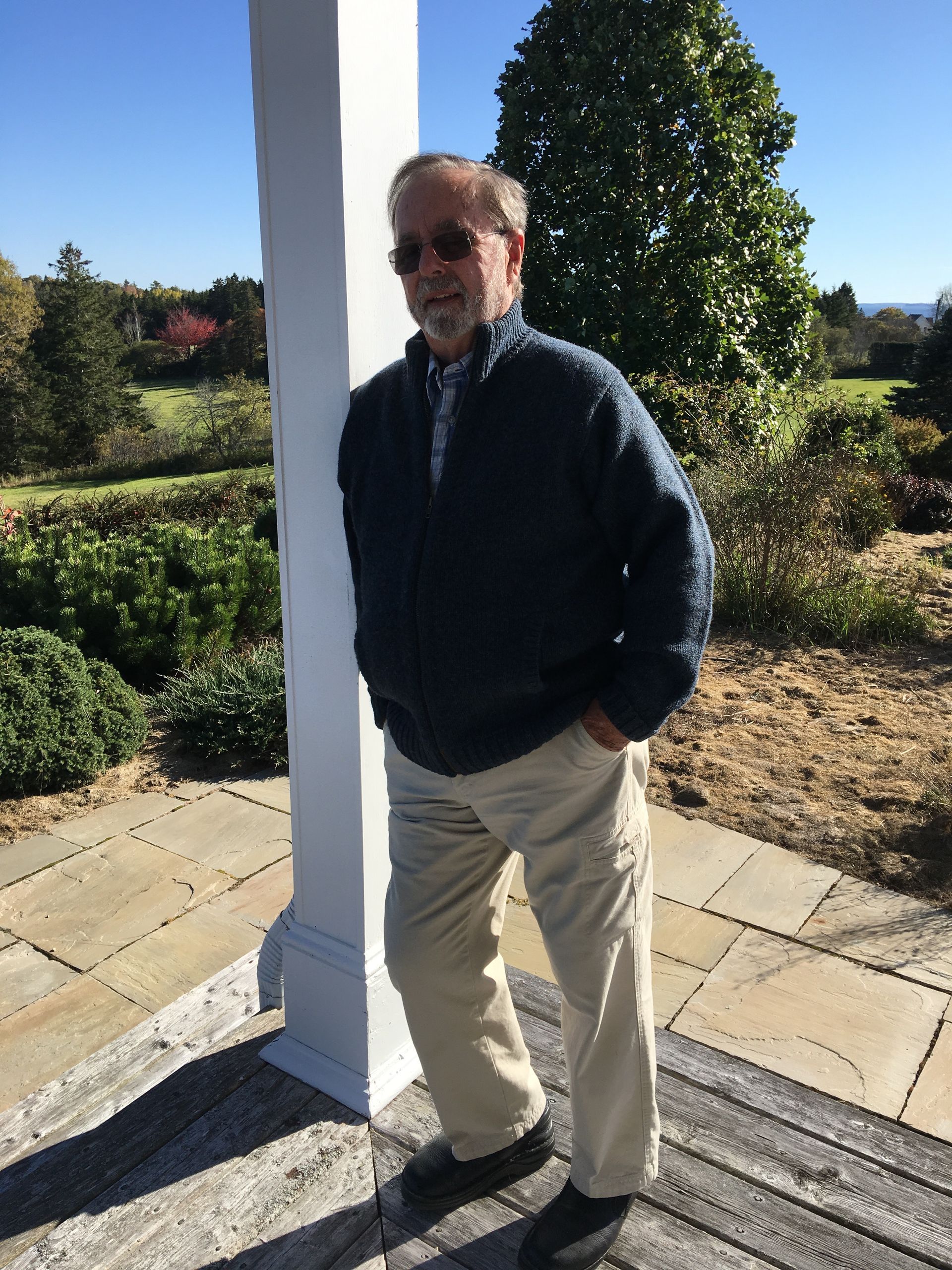 Man in sunglasses and blue sweater leans against a white column outdoors on a sunny day.