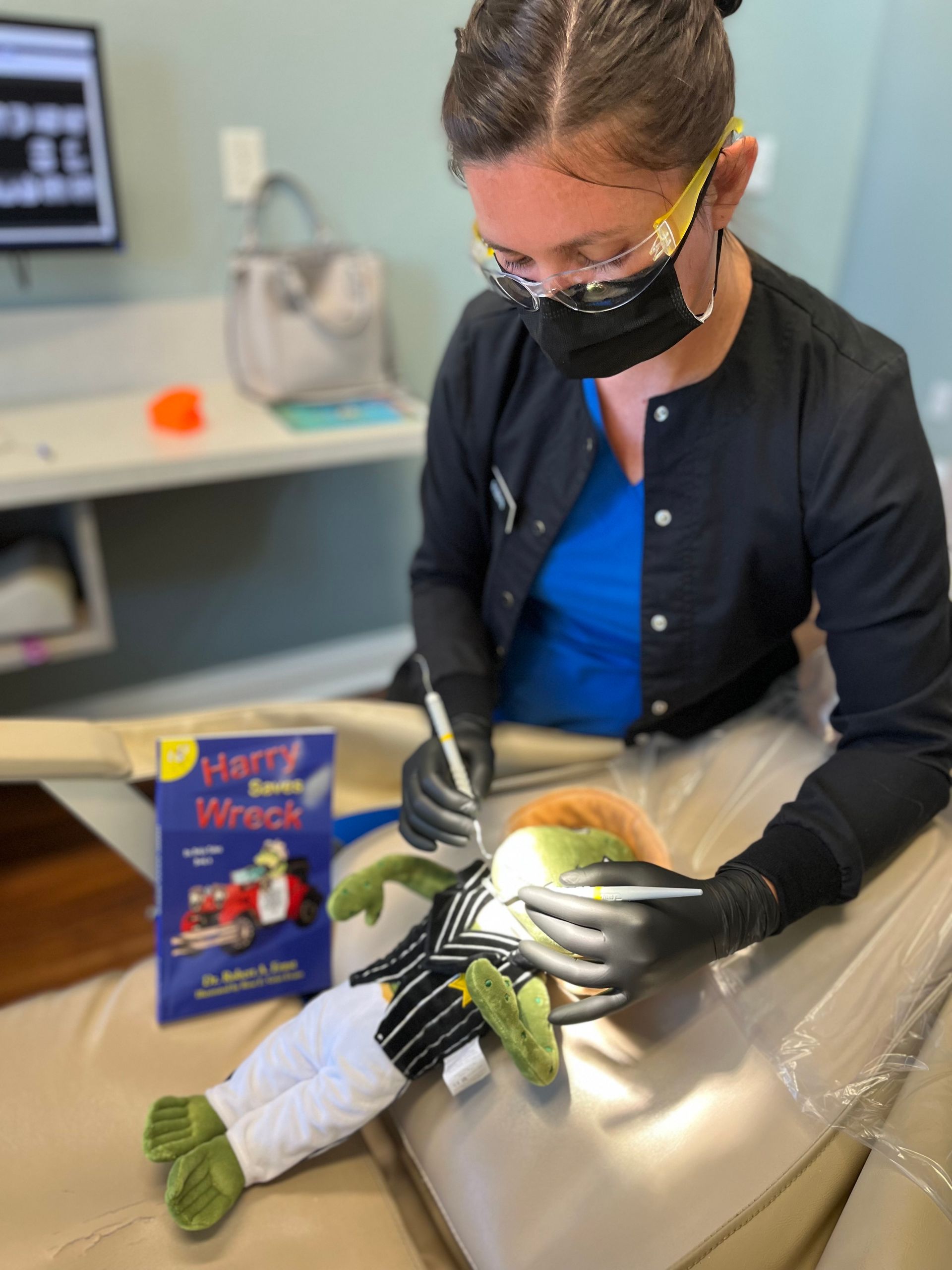 Dentist in black scrubs examines a puppet's teeth with dental tools in a clinic setting. Book visible.