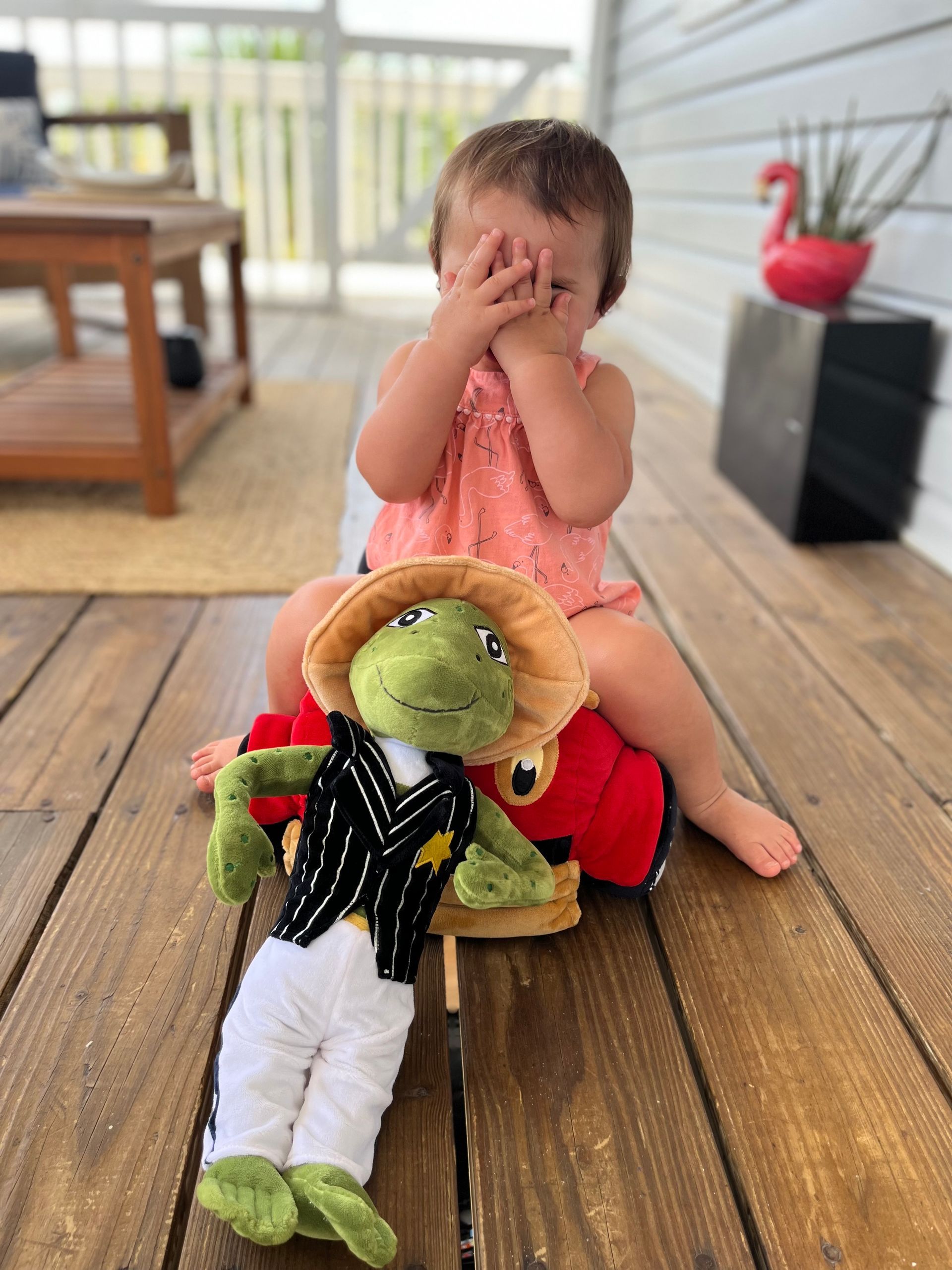 Child covering eyes, sitting with frog toy on wooden porch.