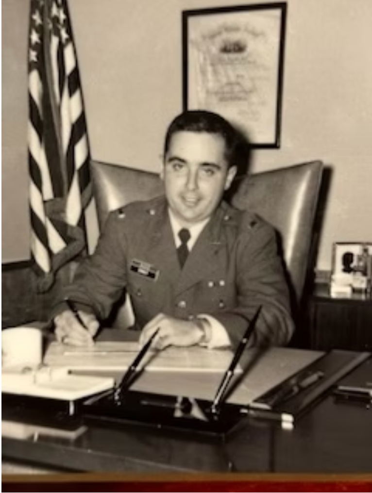 Man in military uniform at desk, writing; US flag and framed document in background.