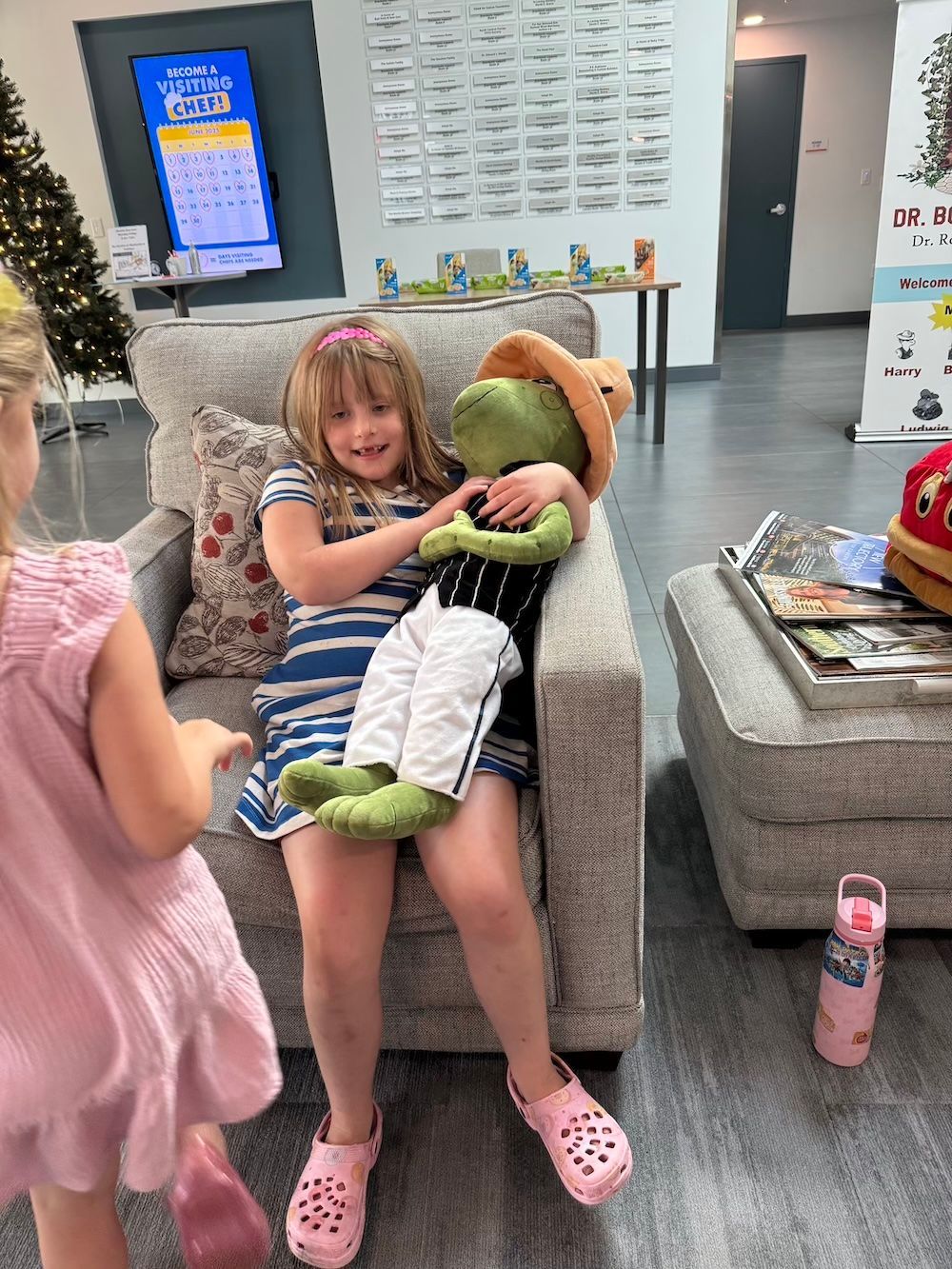 Girl seated, hugging a green stuffed animal with cowboy hat. Another girl nearby; interior waiting area.