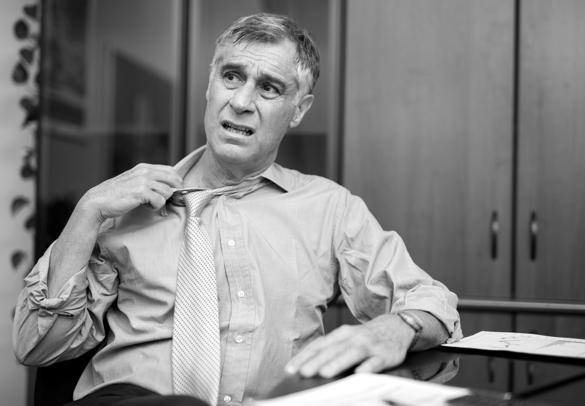 A man is sitting at a desk adjusting his tie. Feeling hot.