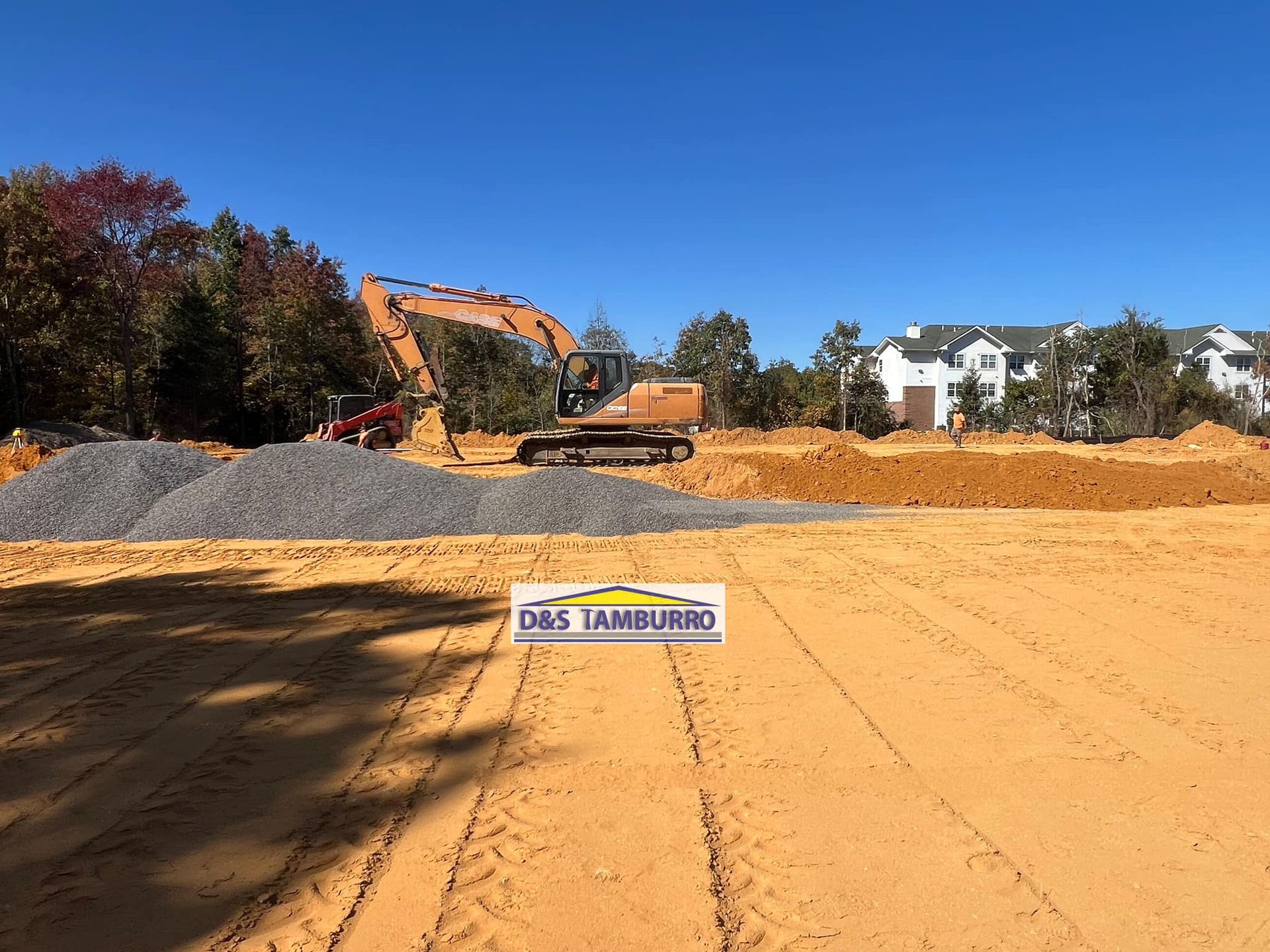 Construction site with excavator and gravel piles under blue sky. Sign reads 