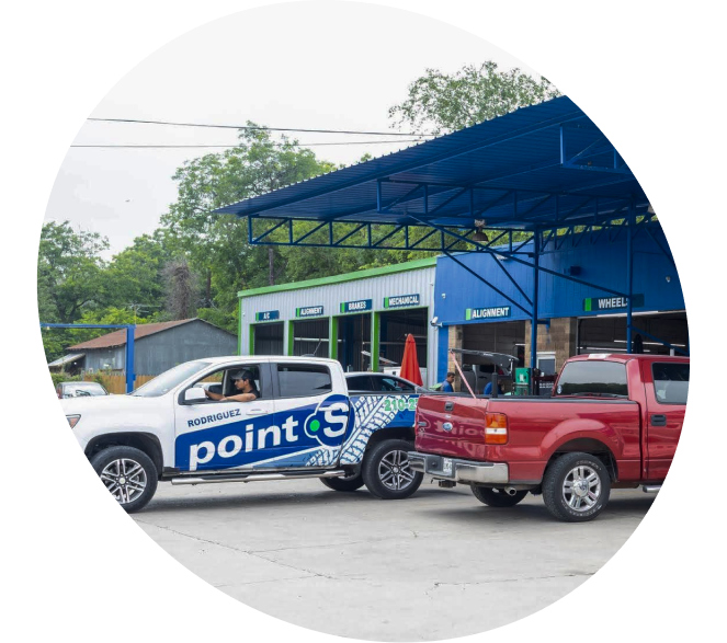 A white branded Point S pickup truck and a red pickup truck parked under a blue metal canopy at an auto repair shop.