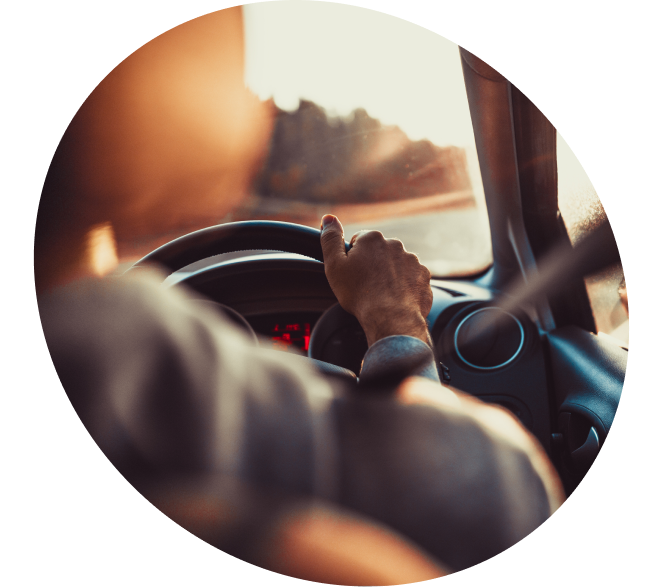 A view from inside a car of a person’s hands on the steering wheel while driving toward a sunset.