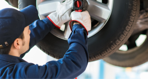 A mechanic in a blue uniform and gloves uses a power tool to tighten a bolt on a car wheel raised in a garage.