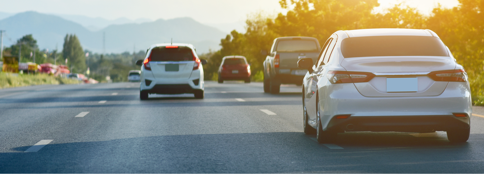 A white car drives in the foreground on a highway with several other vehicles in traffic under sunny skies.