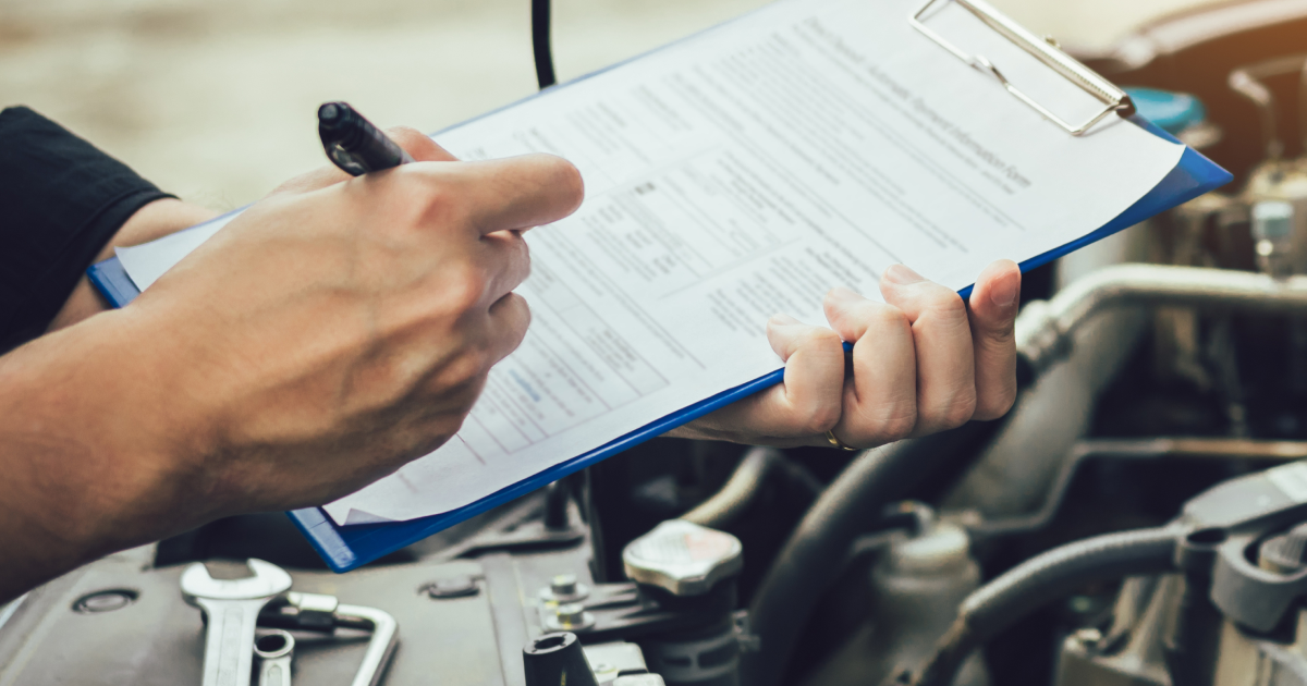 A mechanic’s hands holding a clipboard with a checklist over an open car engine, with tools visible in the foreground.