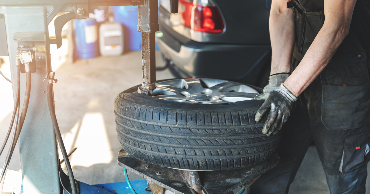 A worker in a shop wearing gloves uses a tire changer machine to service a car tire.