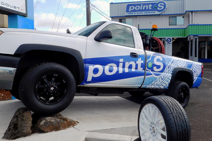 A white pickup truck branded with the blue Point S logo parked on rocks in front of a Point S tire and auto service shop.