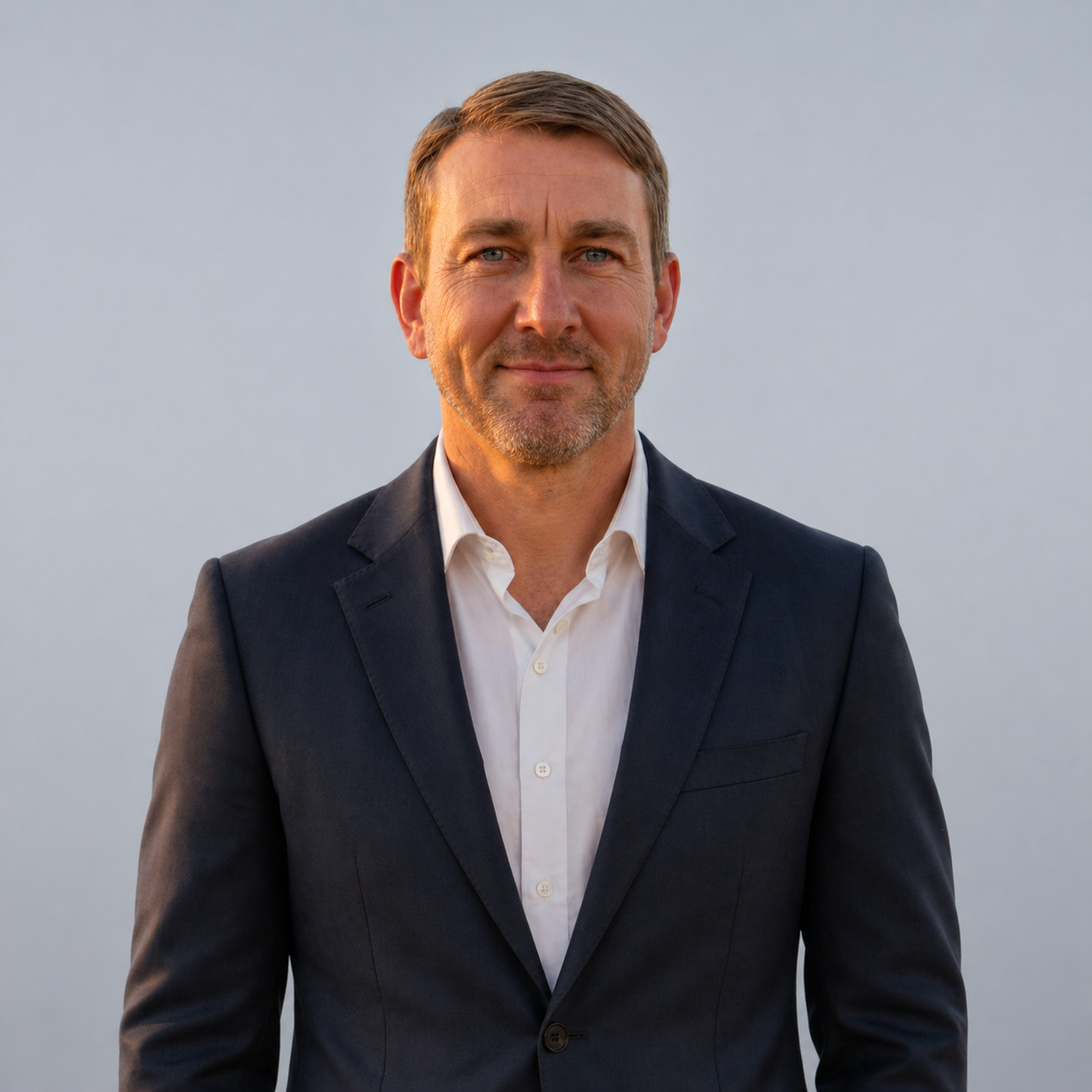 Man in a dark suit and white shirt standing outdoors against a plain gray background.