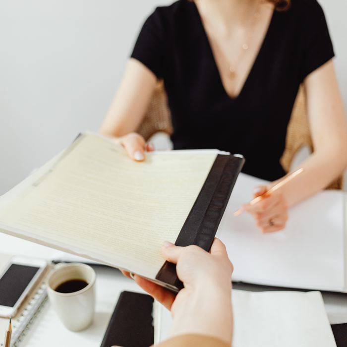 Hands exchanging a black binder at a desk with a coffee cup and laptop, meeting scene
