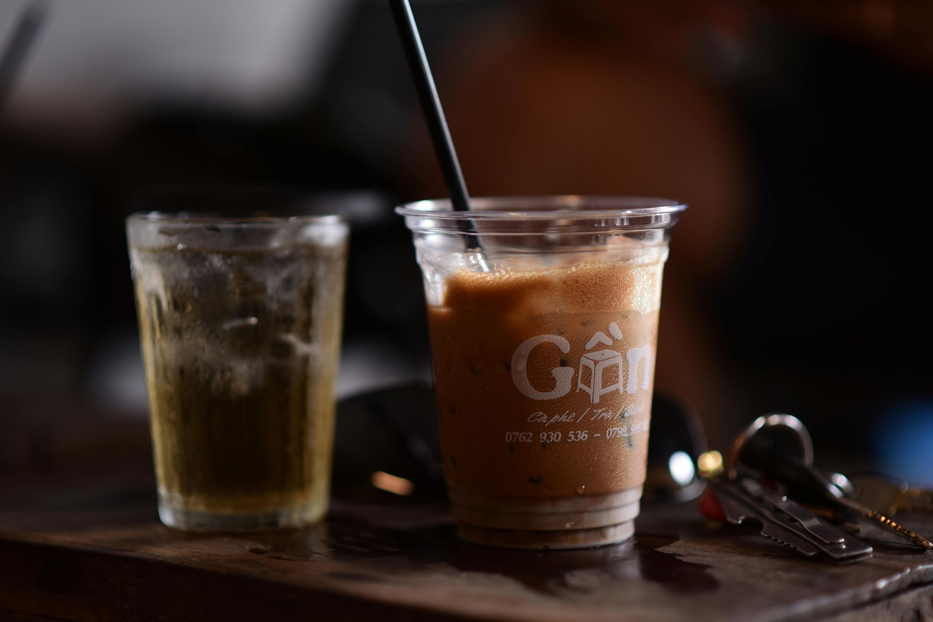 Two iced drinks on a dark table, one in a clear glass and one in a branded takeaway cup with a straw.