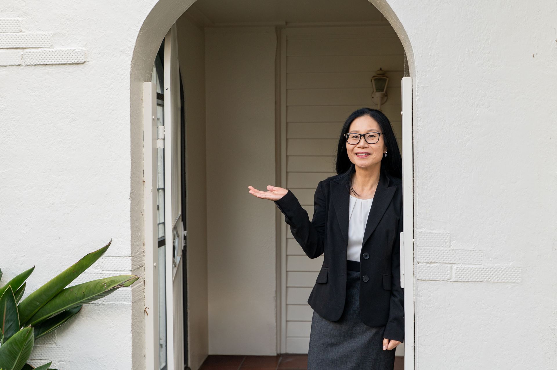 Woman in a black blazer standing in a doorway, gesturing with one hand in a white-walled entryway.