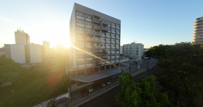 Prédio de apartamentos moderno e ensolarado ao pôr do sol, com árvores e torres vizinhas ao fundo.