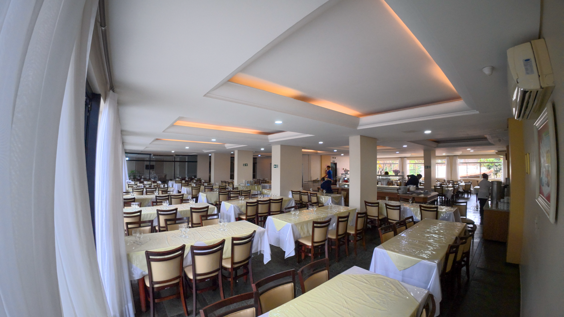 Amplo salão de jantar de restaurante com mesas e cadeiras vazias e iluminação suave no teto.