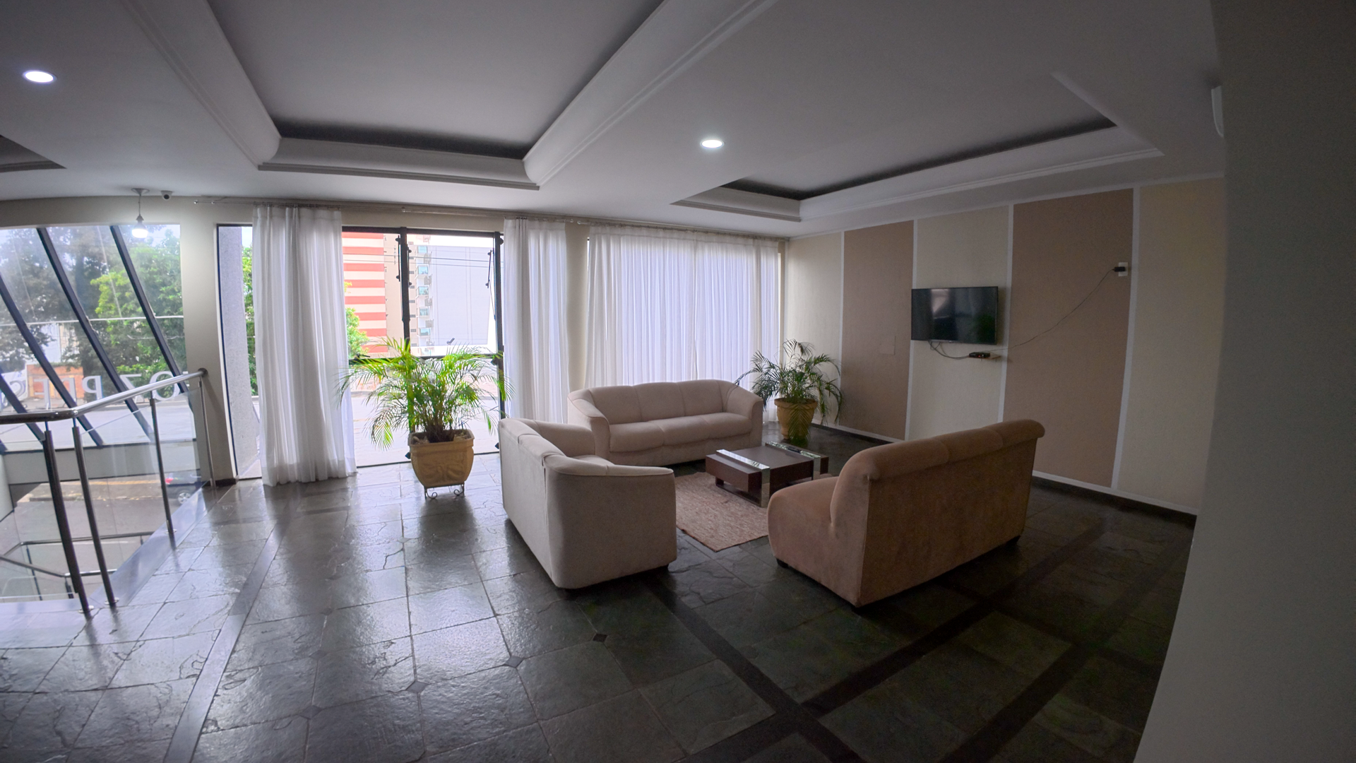 Sala de estar moderna com sofás bege, mesa de centro, janelas amplas e plantas em vasos sobre um piso escuro.