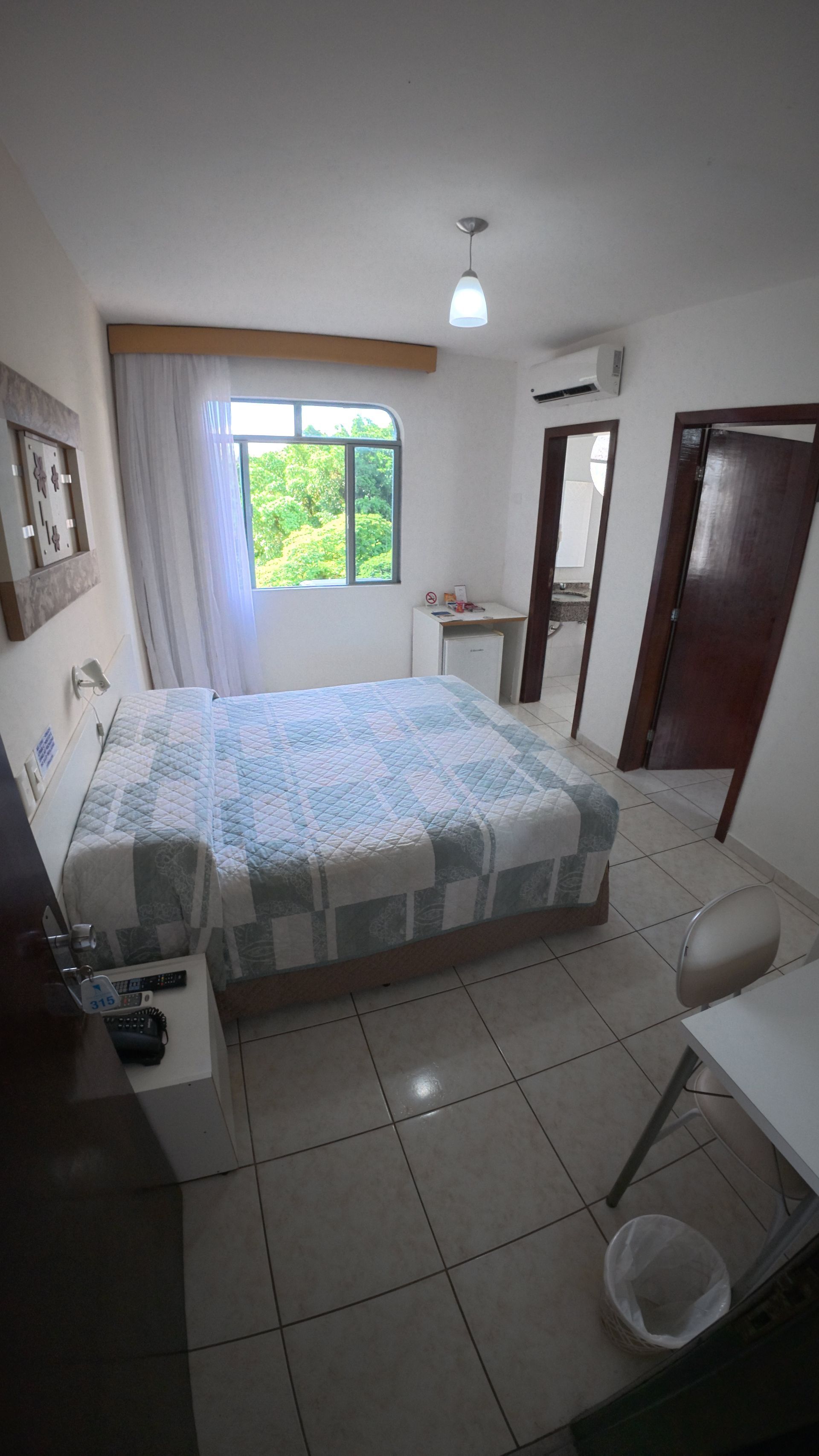 Quarto pequeno com cama, janela, escrivaninha, guarda-roupa, piso de azulejo e paredes brancas.