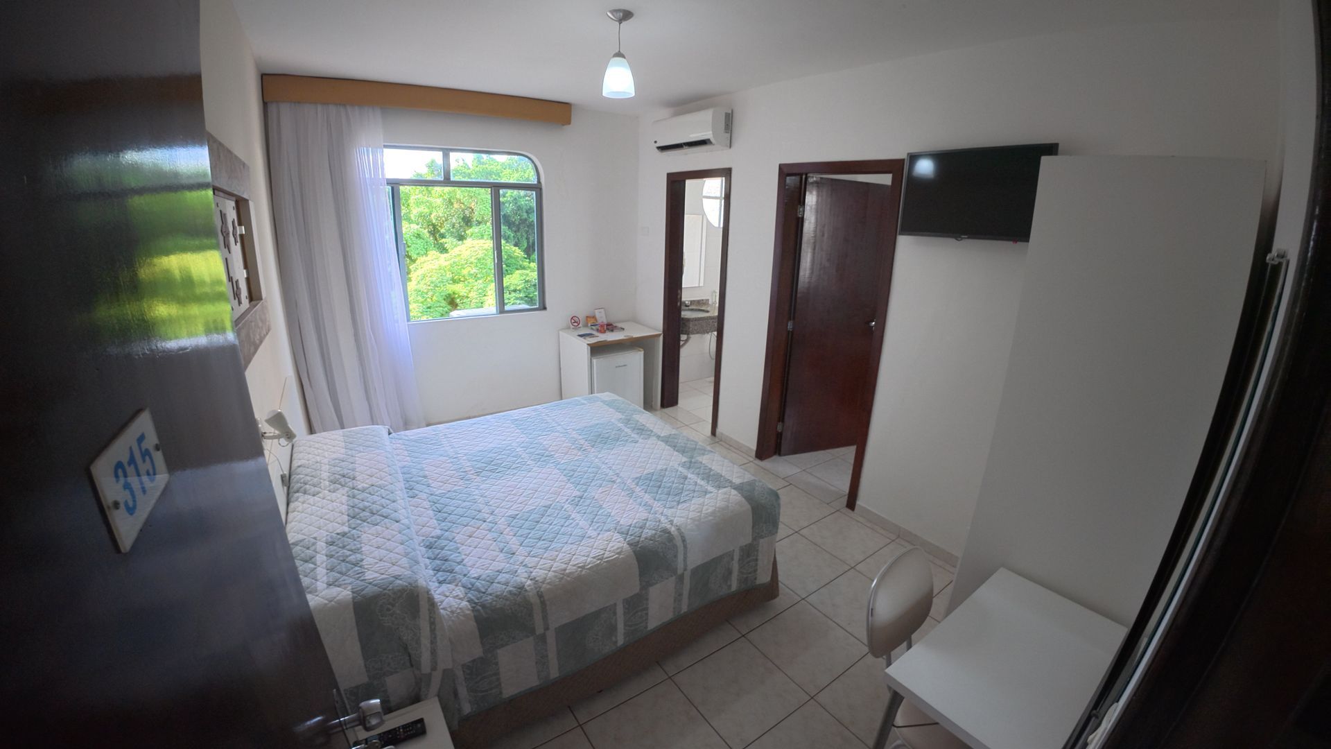 Quarto pequeno com cama, janela, ventilador de teto, TV de parede e piso de azulejo.