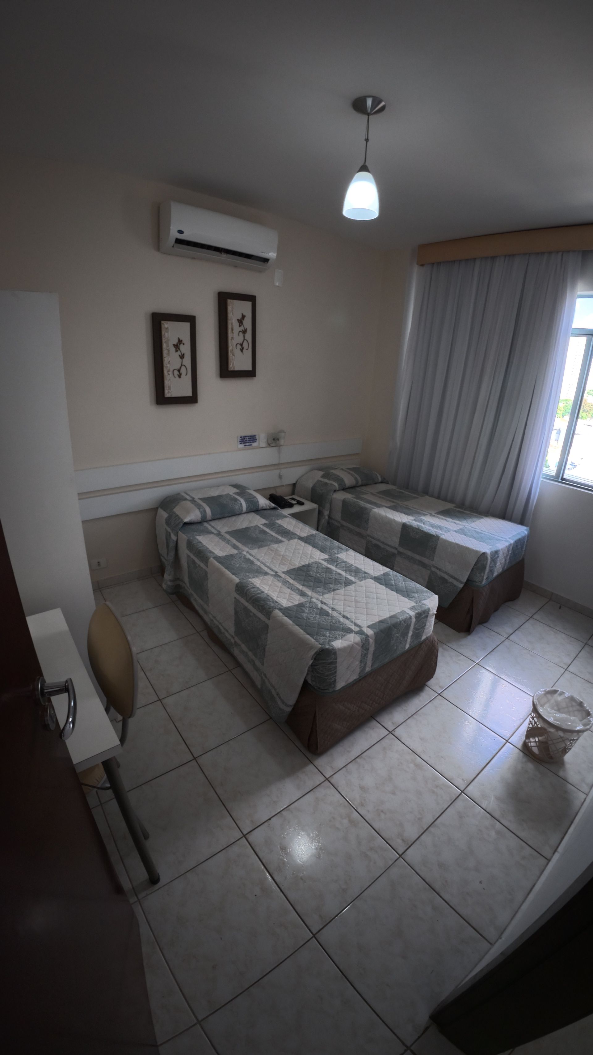 Quarto pequeno com duas camas de solteiro, piso de azulejo, ar-condicionado de parede e janela com cortinas.
