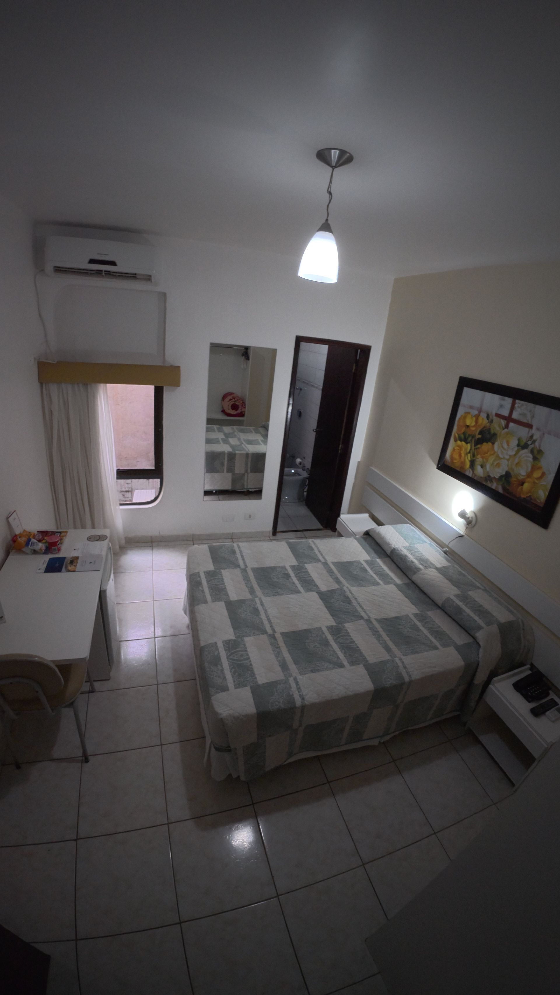 Quarto pequeno com cama, piso de azulejo, luminária pendente, obras de arte na parede e porta aberta para um corredor.