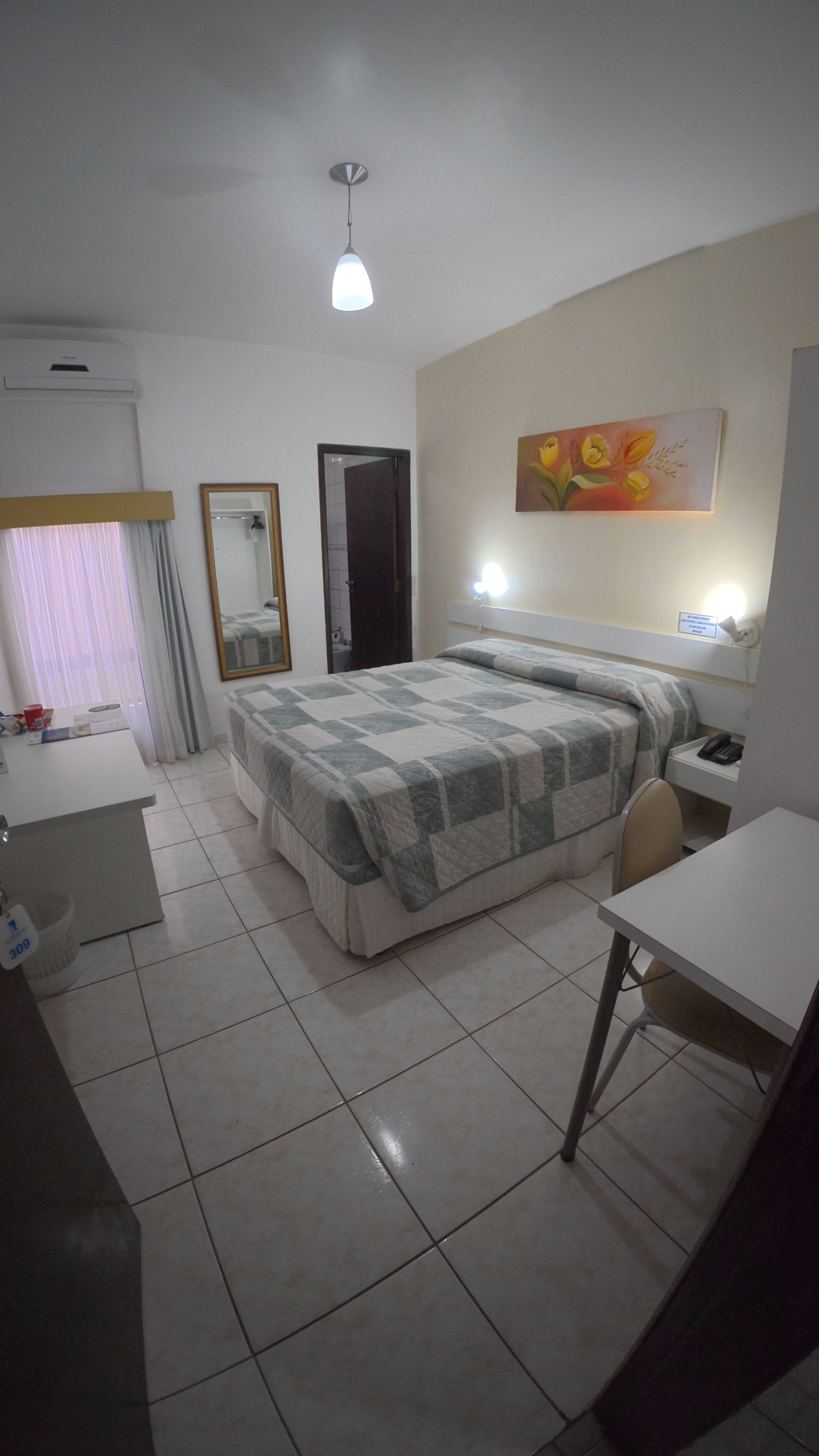 Quarto pequeno com cama, piso de azulejo, obras de arte na parede, espelho e janela com cortinas rosa.