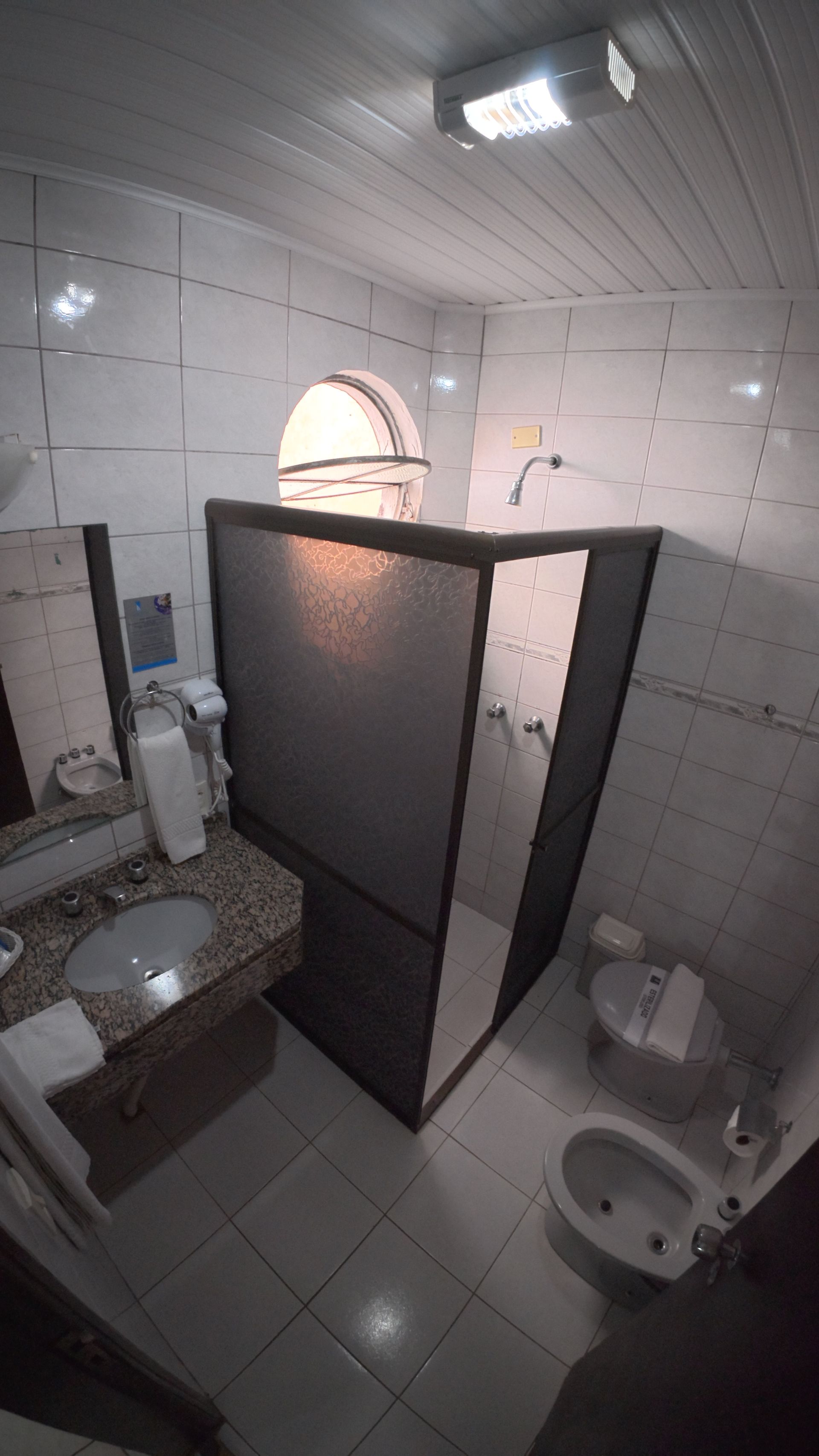 Banheiro público com paredes revestidas de azulejos, box para chuveiro, pia, vaso sanitário e janela fosca.