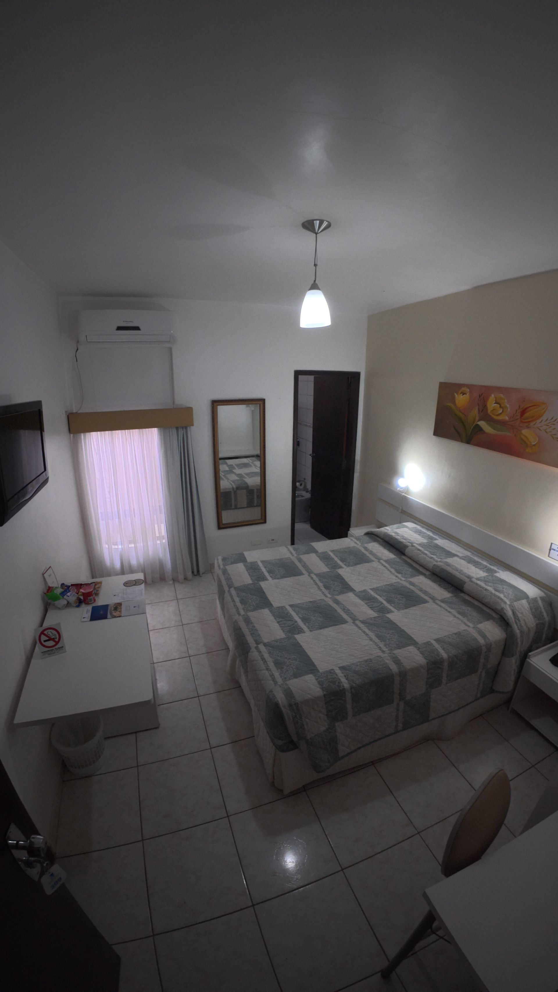 Quarto pequeno com cama, piso de azulejo, obras de arte na parede, espelho e janela com cortinas.