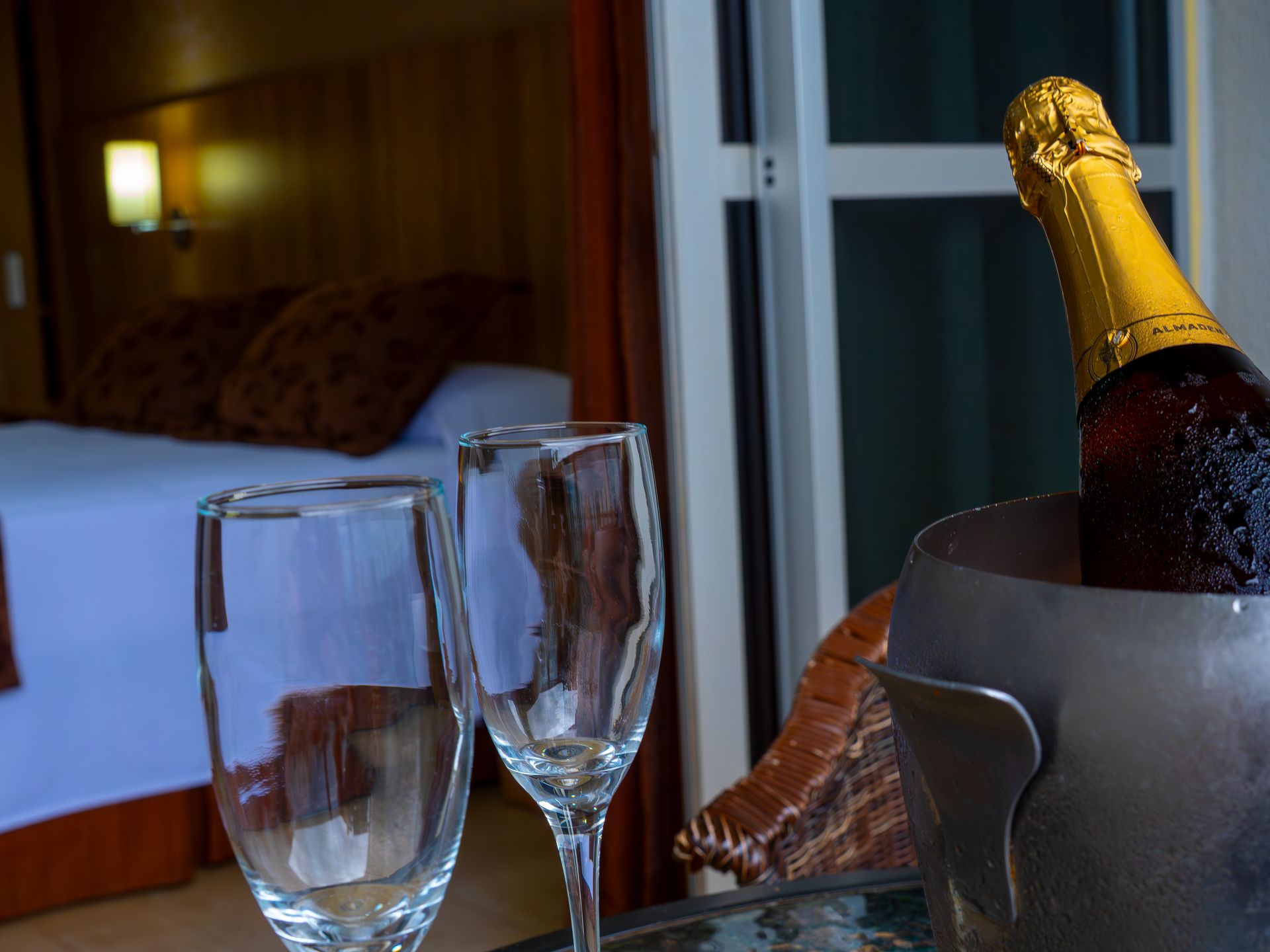 Garrafa de champanhe e taças vazias sobre uma mesa ao lado da cama de um hotel à noite.