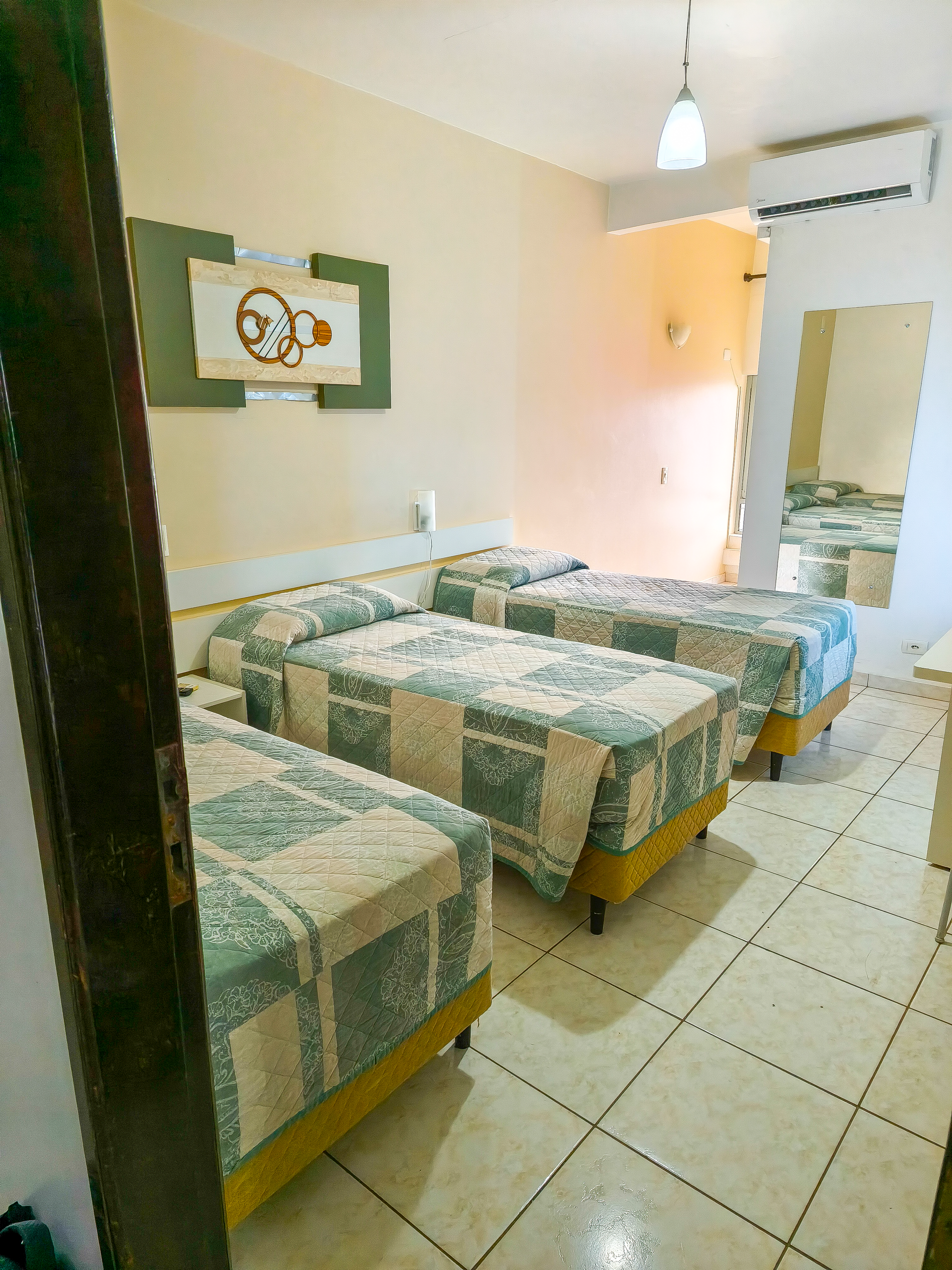 Quarto de hotel com quatro camas de solteiro, piso de azulejo, espelho e porta para um banheiro bem iluminado.