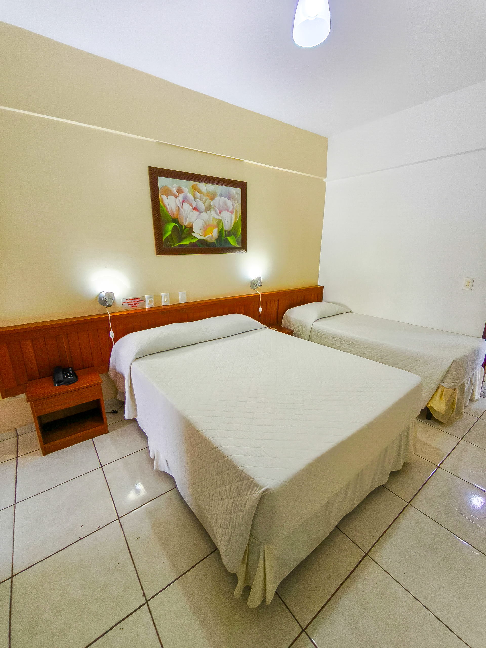 Quarto de hotel pequeno com duas camas, cabeceira de madeira, abajures e piso de azulejo.