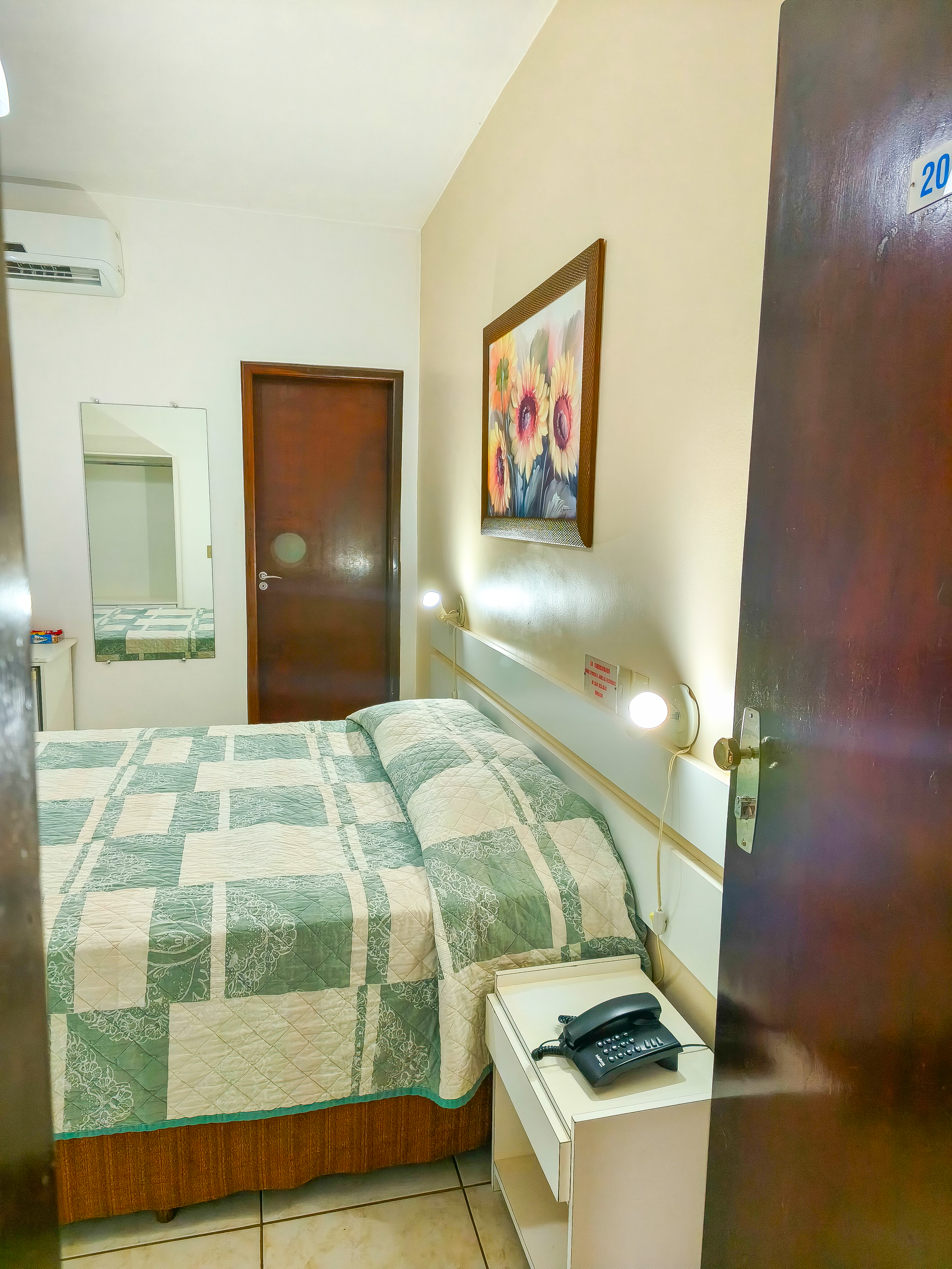 Quarto pequeno com cama de colcha, mesa de cabeceira, obras de arte na parede e porta entreaberta.