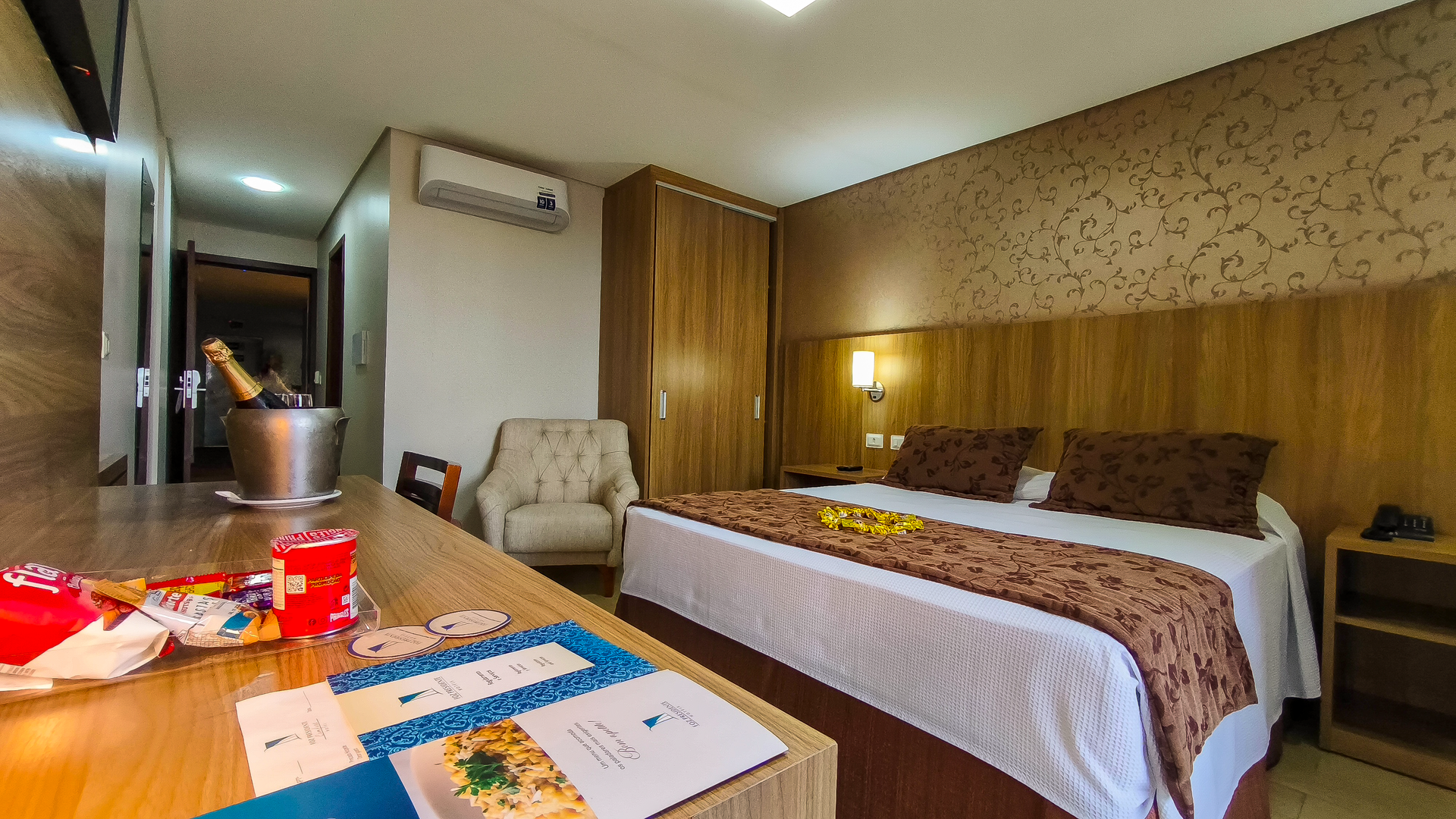 Quarto de hotel moderno com cama bem arrumada, ar condicionado, escrivaninha e decoração aconchegante em madeira.