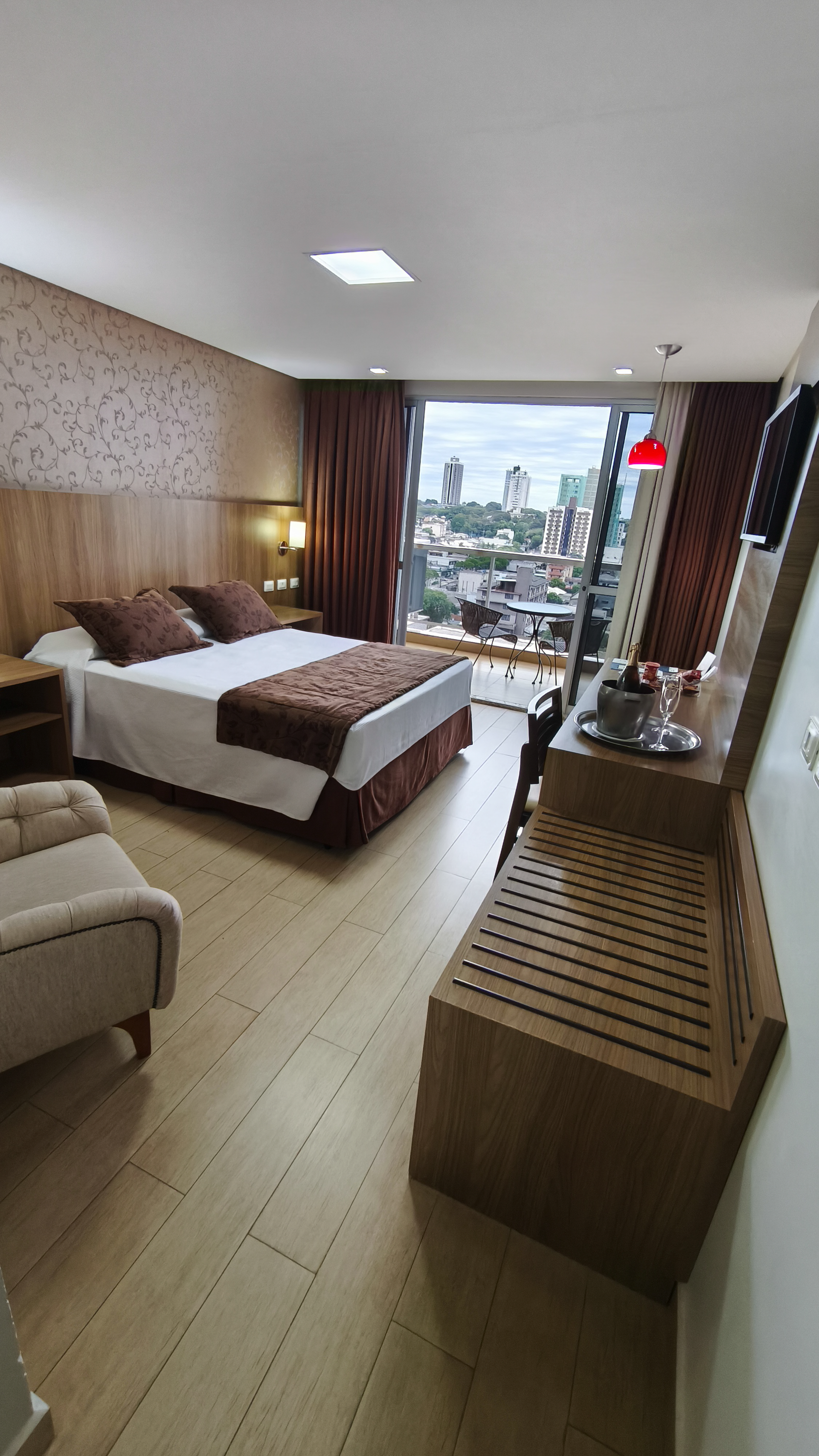 Quarto de hotel com cama, sofá, escrivaninha e janela com varanda com vista para o horizonte da cidade.